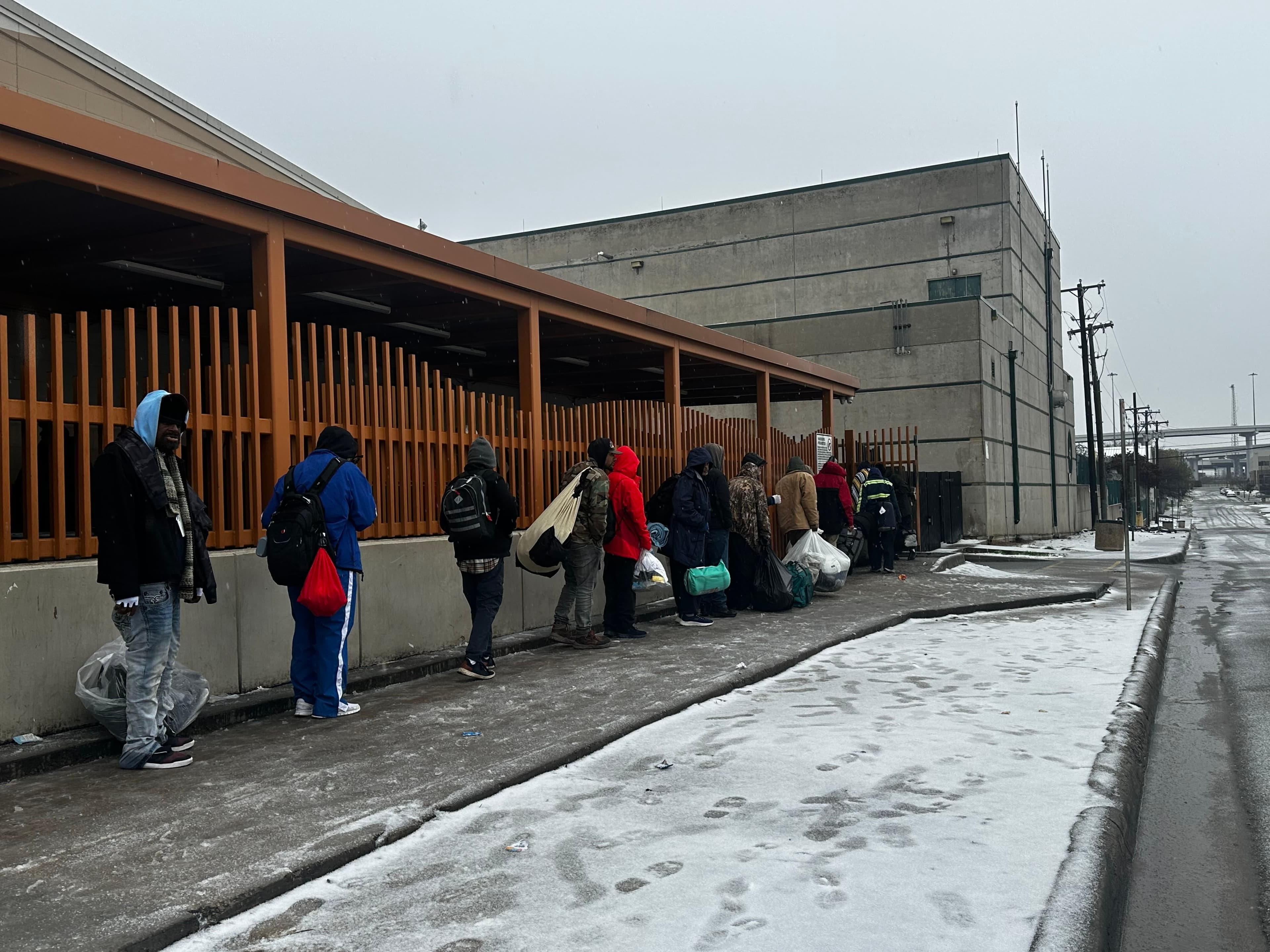 A group of men line up outside of a shelter, snow on the ground.